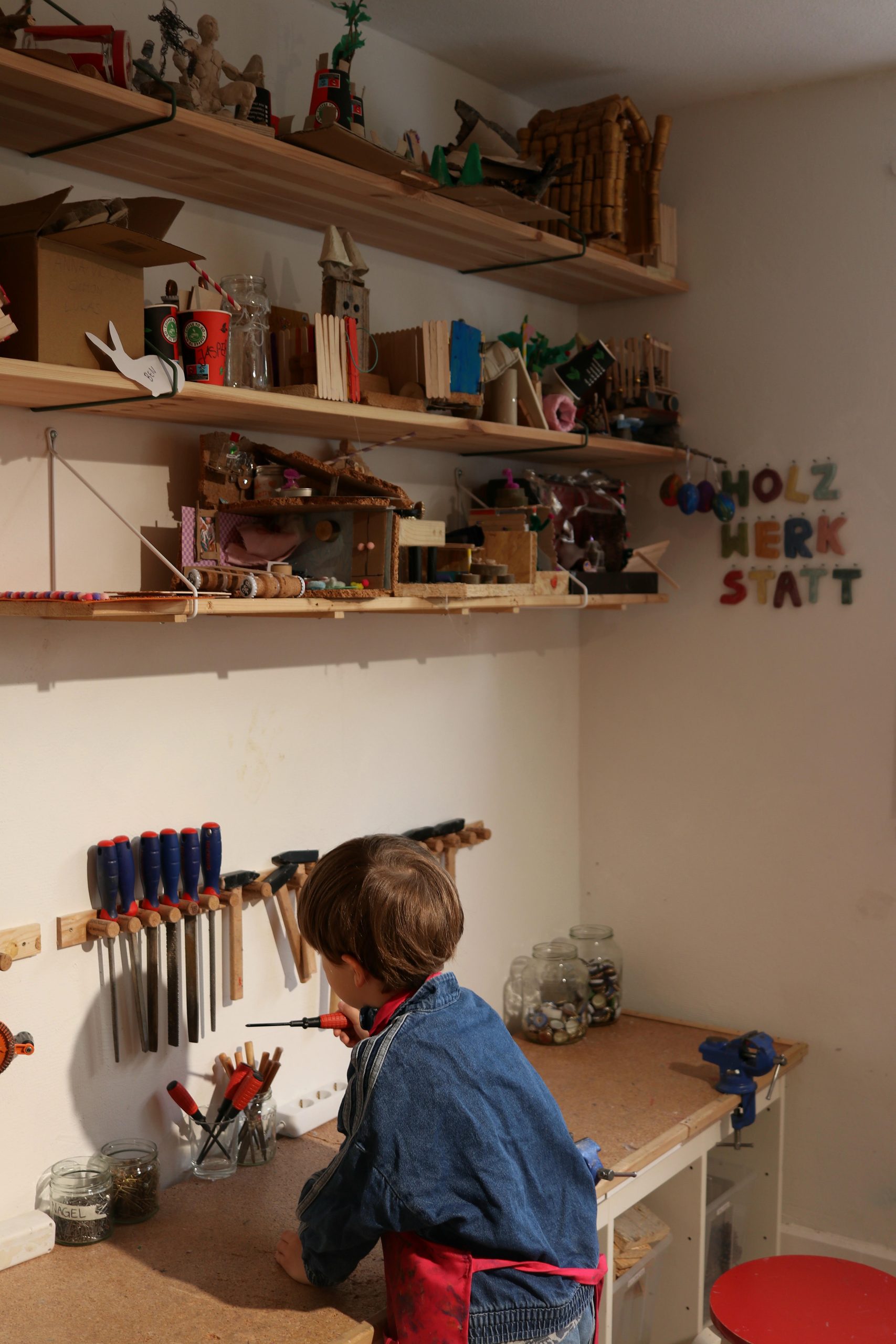 Ein Kind werkelt in der Holzwerkstatt mit einem Schraubenzieher, darüber Regale mit plastischen Kinderkunstwerken und Halter für verschiedene Werkzeuge