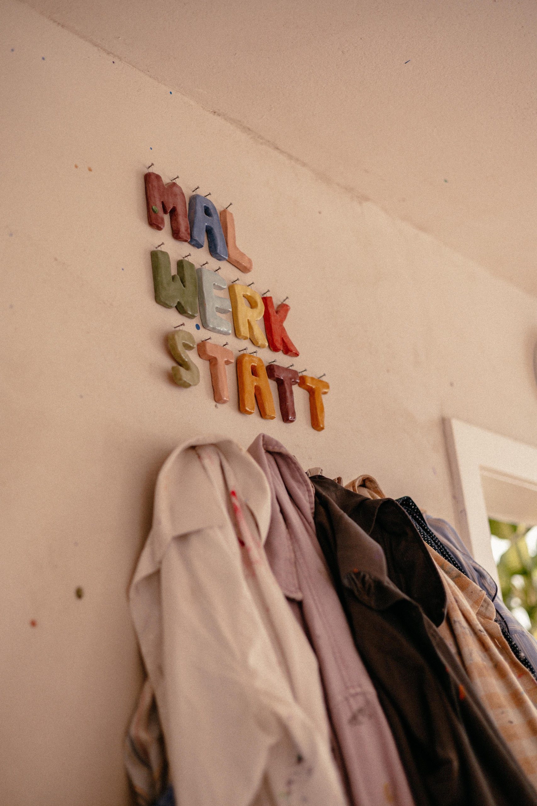 Farbige Tonbuchstaben zeigen das Wort Malwerkstatt, darunter Kunstmäntel für Kinder an Haken.