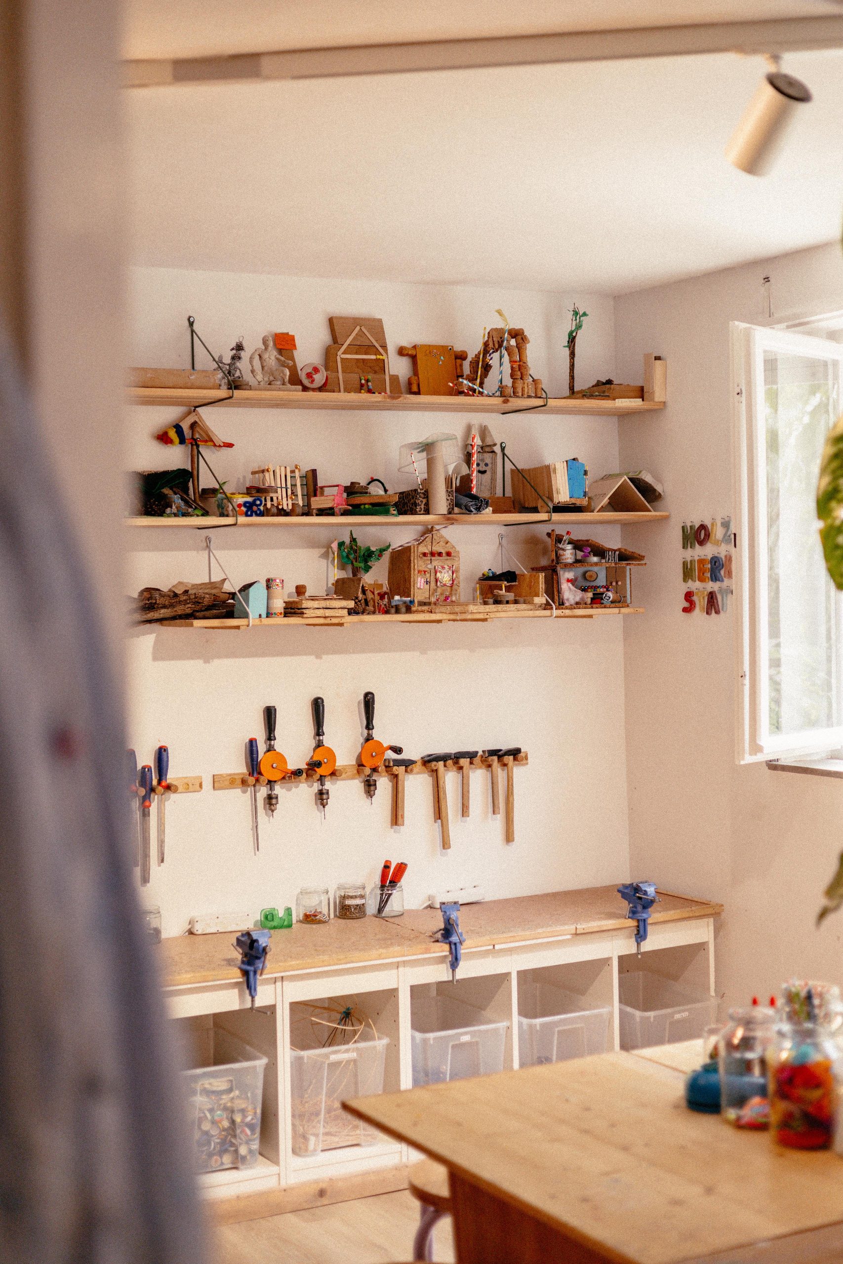 Lichtdurchflutetes Zimmer in der Kindermalwerkstatt mit Regalen voller Kunstwerke der Kinder und darunter Bastelutensilien und Werkzeug